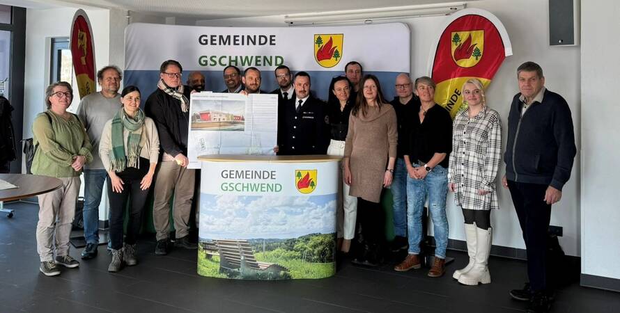 Eine Gruppe von Menschen bestenden aus Vertretern des Gemeinderats, Mitarbeiter des Rathaus Gschwend und Bürgermeister Ziehr steht lächelnd zusammen vor einem Hintergrund mit der Aufschrift "Gemeinde Gschwend" und einer Fahne. Einige halten ein Plakat mit dem Bild des neuen Feuerwehrhaus hoch.