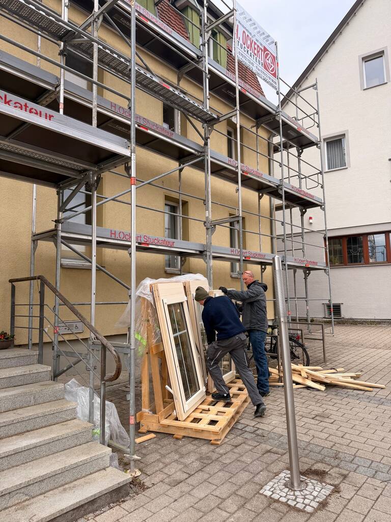 Zwei Männer arbeiten an einem Gebäude mit Gerüst, wobei sie Fensterrahmen auf Holzpaletten untersuchen oder bearbeiten.